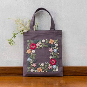 May include: A gray tote bag with embroidered floral design. The embroidery features a square border of colorful flowers, including red, pink, orange, and blue, with green leaves. The bag has two handles and is made of a sturdy fabric.