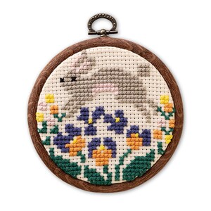 Puede incluir: Un bordado de punto de cruz con un conejito gris con detalles rosas, rodeado de flores azules, amarillas y naranjas. La obra de arte está enmarcada en un marco de madera redondo con un lazo de metal para colgar.