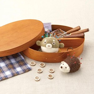 May include: A wooden sewing box with a lid, containing sewing tools and a felt pufferfish pin cushion. A felt hedgehog pin cushion and several wooden buttons are also visible. A plaid fabric is on the left.