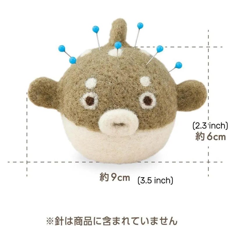 May include: A felt pufferfish pincushion, approximately 3.5 inches wide and 2.3 inches tall. The pincushion is tan and white with blue-tipped pins inserted. The text on the image says "The needles are not included."