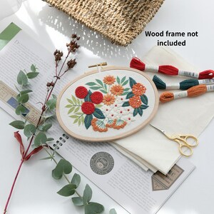 May include: An oval embroidery hoop with a floral design in red, orange, and white. Green leaves and stems surround the flowers. The image includes embroidery floss, fabric, scissors, and the text "Wood frame not included."