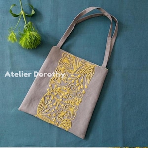 Può includere: Una borsa tote grigia con ricami floreali gialli. La borsa ha due manici grigi e il testo "Atelier Dorothy" sul lato. Il ricamo presenta motivi floreali e fogliari. La borsa è su una superficie blu.
