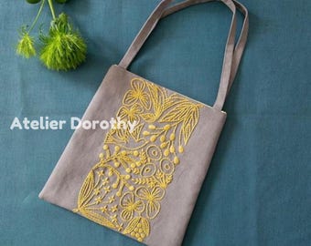 May include: A gray tote bag with yellow floral embroidery. The bag has two gray handles and the text "Atelier Dorothy" on the side. The embroidery features flowers and leaf patterns. The bag is on a blue surface.