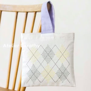 Könnte beinhalten: Eine weiße Tragetasche mit lavendelfarbenem Griff wird auf einem Holzstuhl präsentiert. Die Tasche hat ein gesticktes Rautenmuster in Gelb und Grau. Der Text "Atelier Dorothy" ist auf der Tasche zu sehen.