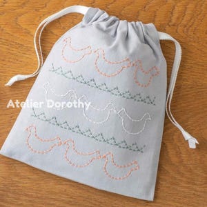 Peut inclure: Un sac en tissu gris clair avec cordon de serrage, orné de broderies de poulets et d'herbes en pêche, blanc et vert. Fermeture par cordon blanc et texte "Atelier Dorothy".