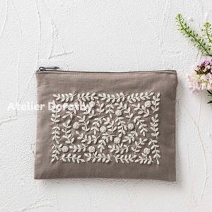 Könnte beinhalten: Eine taupefarbene Reißverschlusstasche mit weißer Blumenstickerei. Die Stickerei zeigt ein Muster aus Blättern und kleinen, runden Blüten. Der Text "Atelier Dorothy" ist auf der linken Seite der Tasche aufgedruckt.