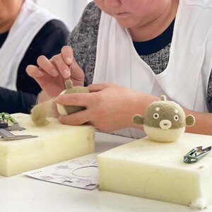 May include: A person is shown crafting a felt pufferfish toy. The toy is primarily beige and white, with small, round eyes and a small mouth. The crafter is using a needle to attach a small piece of felt to the toy. The background is blurred.
