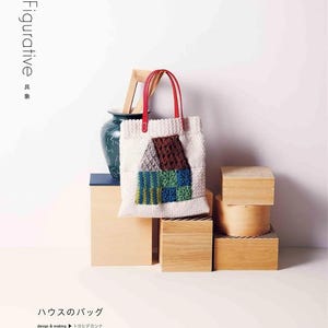Puede incluir: Un bolso tote blanco con asas rojas presenta un dise&ntilde;o de casa de ganchillo en marr&oacute;n, azul y verde. La bolsa se exhibe sobre una pila de cajas de madera, con un jarr&oacute;n decorativo y un marco de madera en el fondo. La palabra "Figurative" est&aacute; impresa.