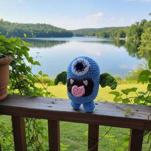 May include: A blue crocheted monster toy with a single large eye, green wings, and a brown mouth with a pink tongue. The handmade plush is on a wooden railing, with a lake and trees in the background.