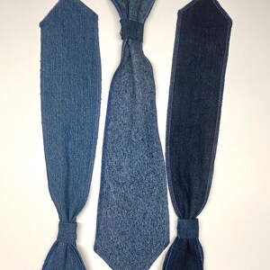 May include: Three denim ties in varying shades of blue are displayed against a white background. The ties have a classic shape, with a knot or band at the neck and a pointed or straight end. The denim fabric has visible texture and stitching.