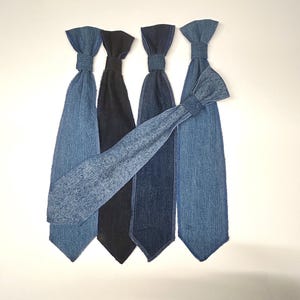 May include: Five denim ties in various shades of blue and black are arranged against a white background. The ties have a classic shape with a knot at the top and a pointed bottom. One tie is angled across the others, showcasing the different denim washes.