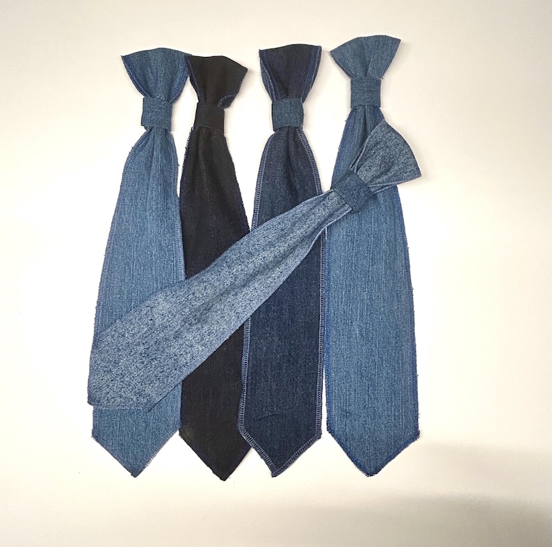 May include: Five denim ties in various shades of blue and black are arranged against a white background. The ties have a classic shape with a knot at the top and a pointed bottom. One tie is angled across the others, showcasing the different denim washes.