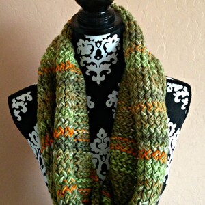 May include: A green and orange crocheted infinity scarf with a textured pattern. The scarf is displayed on a black mannequin with a white floral pattern.