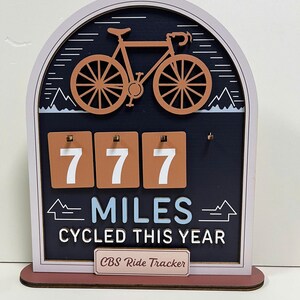 May include: Wooden bicycle ride tracker with a brown bicycle silhouette at the top. The tracker displays "777 MILES CYCLED THIS YEAR" in white and blue lettering. The frame is a light pink color with a brown base.