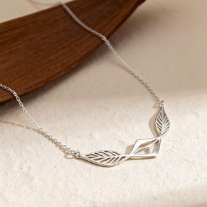 May include: A silver necklace featuring a leaf design. The necklace has a delicate chain and three stylized leaf pendants. The leaves have intricate vein detailing and are arranged in a curved shape. The necklace is displayed on a neutral background.