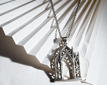 May include: A silver-toned metal pendant shaped like a gothic arched window with intricate tracery and pointed spires, suspended from a matching silver-toned link chain. The pendant features a hollow, open-work design with vertical columns.