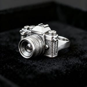May include: A silver-toned ring shaped like a vintage camera. The ring features a detailed lens, a textured grip on the camera body, and various dials on the top. The metal has a polished finish with dark recessed areas to highlight the design.