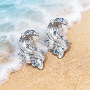 Puede incluir: Un par de pendientes de plata con forma de olas oceánicas estilizadas, con un acabado metálico pulido, pequeñas perlas blancas incrustadas y diminutas piedras preciosas azules redondas dispersas por las crestas curvas y fluidas.