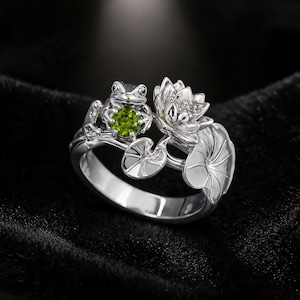 Puede incluir: Un anillo de plata que presenta una rana esculpida sosteniendo una piedra preciosa verde redonda y facetada. La banda está decorada con motivos detallados de flor de loto y nenúfar en plata, creando un diseño coherente inspirado en la naturaleza.