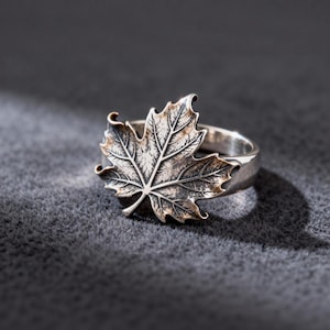 Puede incluir: Anillo de plata con un diseño detallado de hoja de arce. El anillo tiene un acabado texturizado y oxidado, con las venas de la hoja claramente visibles. El anillo se encuentra sobre un fondo gris oscuro, resaltando los intrincados detalles de la joya.