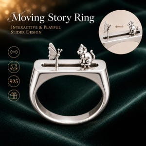May include: A silver ring featuring a horizontal slider track on the top band. A small silver butterfly and a silver cat are positioned on the track. The text Moving Story Ring, Interactive & Playful Slider Design, and 925 are visible.