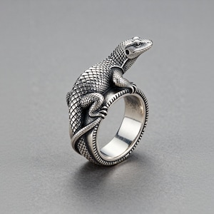 May include: A silver ring with a detailed lizard design. The lizard has textured scales and wraps around the ring band. The ring has a polished finish. The ring is set against a grey background. Jewellery.