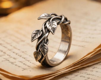 May include: A silver-toned metal ring featuring a band designed with intertwined vines and multiple small, detailed leaves. The surface of the metal has a darkened, oxidized finish that highlights the texture of the foliage and the vine-like structure.