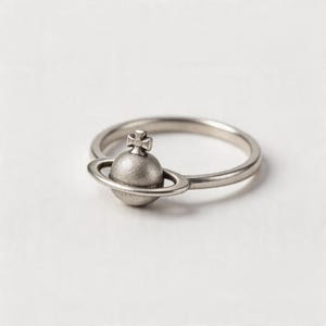 Può includere: Un anello in metallo color argento con un ornamento centrale a forma di pianeta circondato da un anello e sormontato da una piccola croce. La fascia è sottile e circolare, con una finitura opaca e testurizzata sulla sfera planetaria.