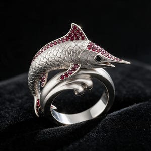 May include: A silver ring featuring a sculpted fish design with a textured scale pattern. The fish is adorned with numerous small, deep red gemstones along its dorsal fin, head, and tail. The ring band is shaped like a smooth, stylized wave.