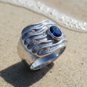 Puede incluir: Un anillo de plata con una gran piedra preciosa ovalada de color azul oscuro en el centro. El anillo tiene un diseño ondulado con pequeñas piedras de acento transparentes. El anillo está sobre una superficie arenosa con olas oceánicas al fondo.