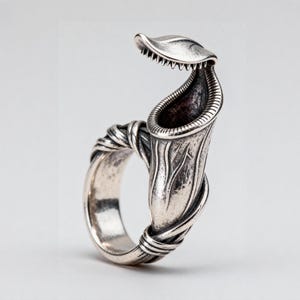 Puede incluir: Un anillo de plata con un diseño tridimensional de planta carnívora. La banda está formada por hilos de metal texturizados que parecen enredaderas que se envuelven alrededor del dedo, culminando en la estructura detallada de la planta abierta.