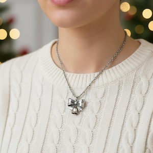 Puede incluir: Un collar de tono plateado con un colgante en forma de lazo y una peque&ntilde;a campana que cuelga del centro. El lazo tiene un acabado met&aacute;lico texturizado con peque&ntilde;os recortes en forma de estrella. La cadena es de eslabones met&aacute;licos sencillos.