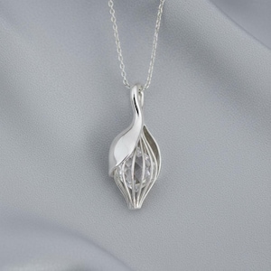 Puede incluir: Un collar con colgante de plata con un diseño en forma de jaula que contiene una piedra clara y facetada. El colgante cuelga de una fina cadena de plata. El fondo es una tela gris suave.