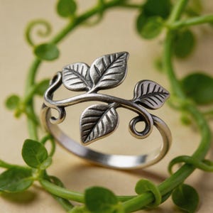 May include: A silver-toned metal ring featuring a band that transitions into a decorative branch motif. The design includes three detailed leaves with etched veins and two small curled tendrils extending from the central vine structure.