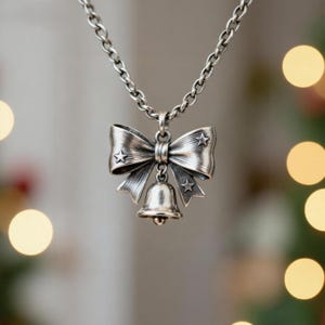 Puede incluir: Un collar de tono plateado con un colgante en forma de lazo decorado con tres peque&ntilde;as estrellas en relieve. Una peque&ntilde;a campana cuelga del centro del lazo. El colgante est&aacute; unido a una cadena de metal plateado con eslabones redondeados.