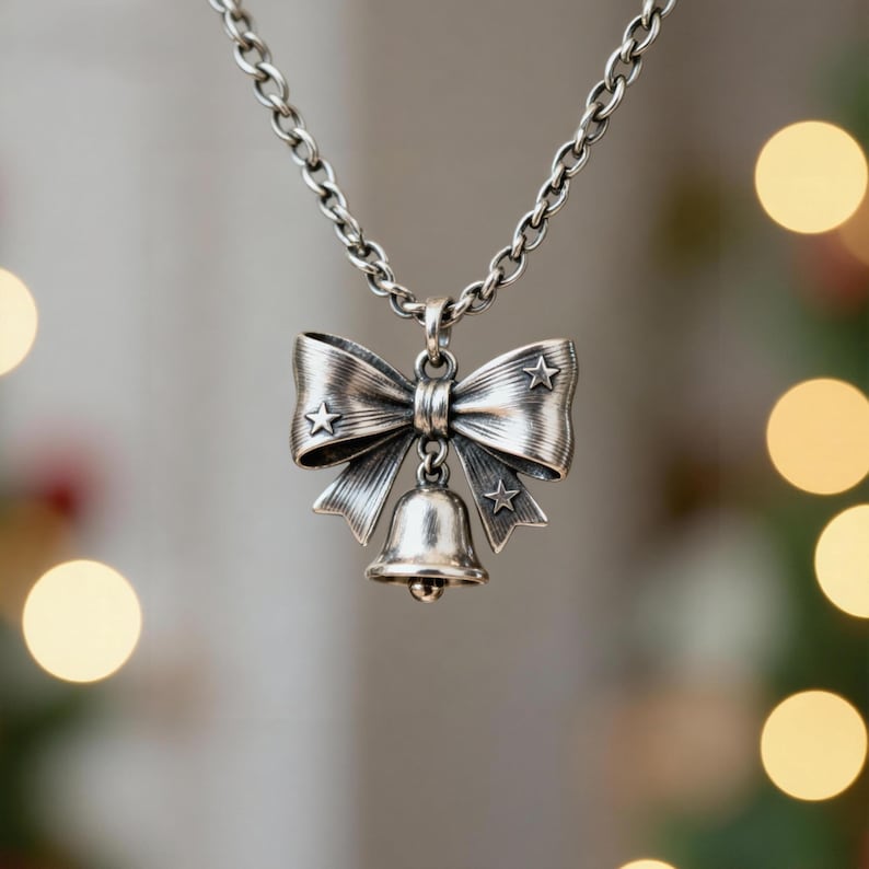 Puede incluir: Un collar de tono plateado con un colgante en forma de lazo decorado con tres peque&ntilde;as estrellas en relieve. Una peque&ntilde;a campana cuelga del centro del lazo. El colgante est&aacute; unido a una cadena de metal plateado con eslabones redondeados.