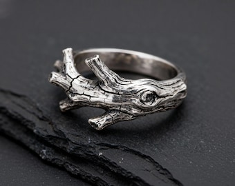 Può includere: Un anello in argento caratterizzato da un design dettagliato e testurizzato che imita l'aspetto di un ramo d'albero nodoso. La superficie metallica presenta solchi profondi, crepe e nodi, con una finitura lucida che esalta i motivi della corteccia.