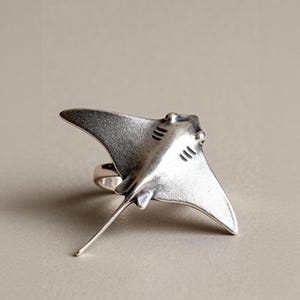 May include: A silver-coloured ring shaped like a manta ray. The ring has a detailed design of the ray's body, including its wings, head, and tail. The ring is set against a neutral background.