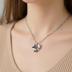 Puede incluir: Un collar de tono plateado con un colgante en forma de lazo que tiene una peque&ntilde;a campana colgando del centro. El metal tiene un acabado oxidado con peque&ntilde;os recortes en forma de estrella en el lazo, suspendido de una cadena de eslabones.