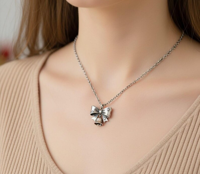 Puede incluir: Un collar de tono plateado con un colgante en forma de lazo que tiene una peque&ntilde;a campana colgando del centro. El metal tiene un acabado oxidado con peque&ntilde;os recortes en forma de estrella en el lazo, suspendido de una cadena de eslabones.