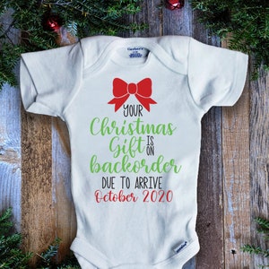 Könnte beinhalten: Weißer Baby-Strampler mit einer roten Schleife und grünem Text, der lautet: "Dein Weihnachtsgeschenk ist auf Vorbestellung und wird voraussichtlich im Oktober 2020 eintreffen."