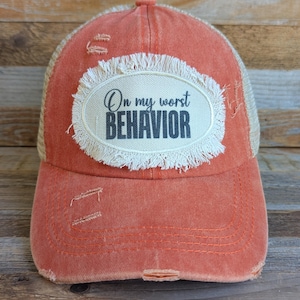 May include: A distressed orange baseball cap with a mesh back and a frayed-edge oval patch on the front. The patch features the black text On my worst BEHAVIOR. The cap has a curved brim and visible stitching details across the front panel.