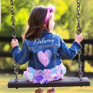 Puede incluir: Una chaqueta vaquera azul con un diseño de corazón y el nombre "Delaney" en escritura blanca. La chaqueta tiene un dobladillo con volantes con detalles de tela rosa y morada. La niña está en un columpio.