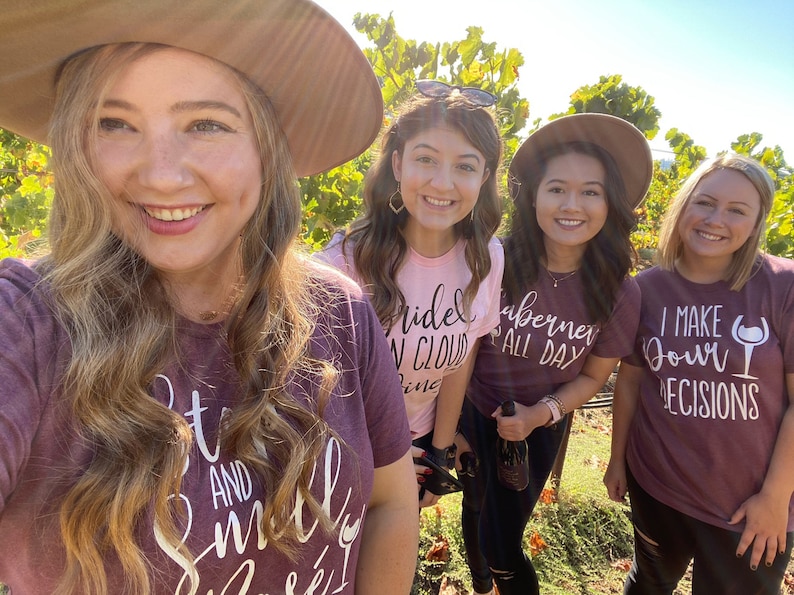 May include: Four friends wearing maroon and pink t-shirts with wine-themed text. The text on the shirts reads "Bride in Cloud Nine", "Cabernet All Day", "I Make My Own Decisions", and "Sit and Smell the Wine".