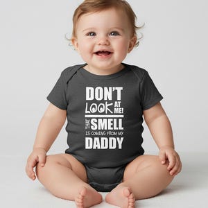 Puede incluir: Un bebé sonriente con un body gris oscuro con texto blanco que dice "DON'T LOOK AT ME! THAT SMELL IS COMING FROM MY DADDY". El bebé está sentado y mirando a la cámara.