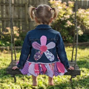 May include: A dark blue denim jacket featuring a large, multi-patterned fabric flower appliqué on the back. The jacket has a ruffled, layered skirt hem made from various shades of purple and pink patterned fabrics, including gingham and floral prints.