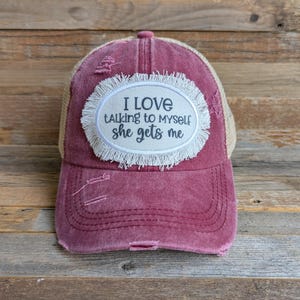 May include: A distressed burgundy baseball cap with a tan mesh back. The cap features an oval patch with the text "I LOVE talking to myself she gets me" in black lettering, surrounded by a white fringe.