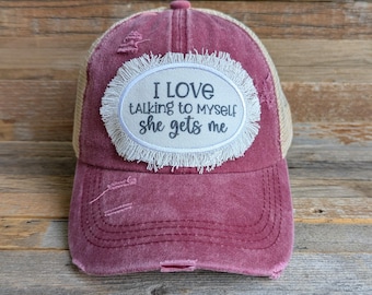 May include: A distressed burgundy baseball cap with a tan mesh back. The cap features an oval patch with the text "I LOVE talking to myself she gets me" in black lettering, surrounded by a white fringe.
