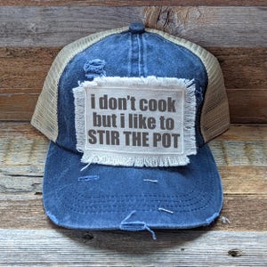 May include: A blue and beige trucker hat with a distressed denim front and a mesh back. The hat has a white patch with the text "I don't cook but I like to stir the pot" in black.