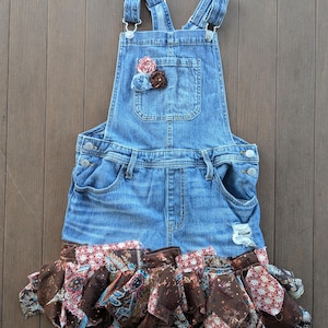 Puede incluir: Un vestido con peto de mezclilla con una falda deshilachada y un adorno floral decorativo en el bolsillo del pecho. La falda presenta un diseño con volantes con tela estampada en tonos marrones, rojos y azules.